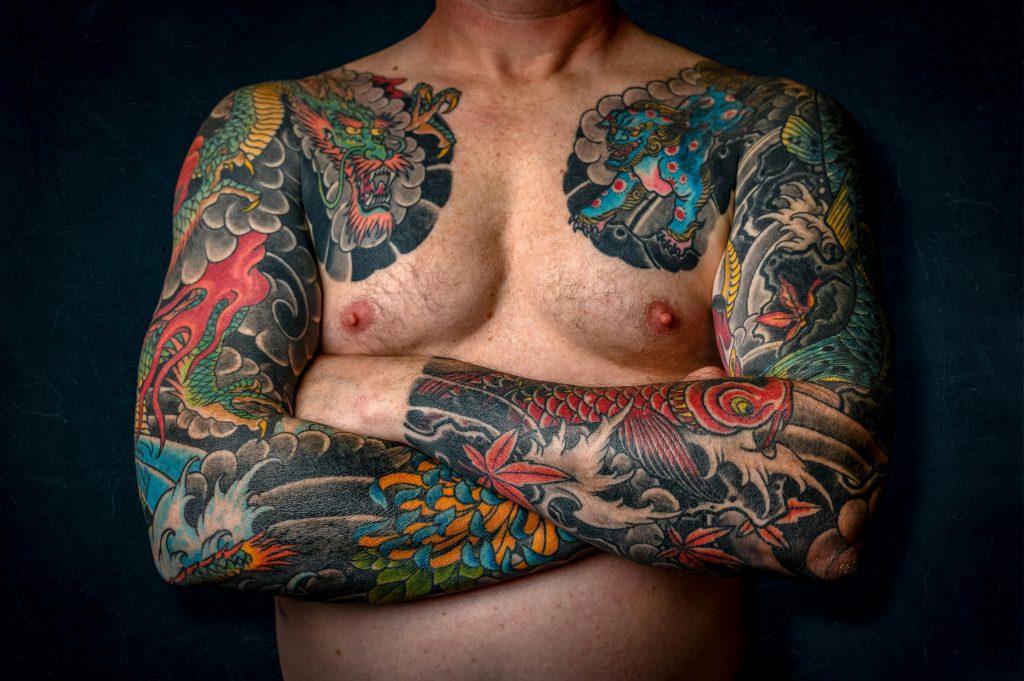 Close-up of a shirtless man with colorful sleeve tattoos and chest art.
